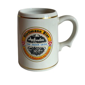 May include: White ceramic beer stein with a gold rim and handle. The stein features a circular emblem with the words "Hürlimann Bier" and "Brauerei A. Hürlimann a/g Zürich" around a central image of a building and figures.