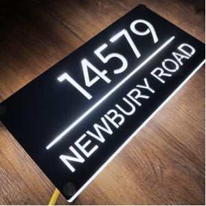 Może przedstawiać: Prostokątny czarny szyld adresowy z podświetlanymi białymi cyframi i tekstem. Cyfry "14579" znajdują się nad tekstem "NEWBURY ROAD". Znak jest oświetlony jasnym białym światłem.