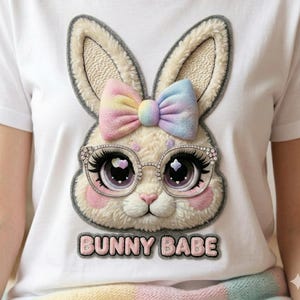 Peut inclure: T-shirt blanc avec un motif de lapin de dessin animé. Le lapin porte des lunettes et un nœud aux couleurs de l'arc-en-ciel pastel. Le texte "BUNNY BABE" est en dessous. Le visage du lapin est détaillé avec de grands yeux et des joues roses.