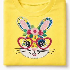 Peut inclure: T-shirt jaune plié avec un visage de lapin brodé. Le lapin porte des lunettes roses en forme de cœur, des oreilles fleuries et une couronne florale. Le motif est orné d'une bordure perlée blanche et de moustaches noires.