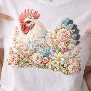 Peut inclure: T-shirt blanc avec une illustration détaillée d'une poule entourée de fleurs. La poule a une crête et des barbillons rouges, avec des plumes bleues et grises. Les fleurs sont roses, blanches et jaunes.