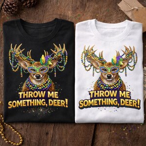 May include: Two t-shirts, one black and one white, featuring a deer graphic with Mardi Gras beads and a mask. The text "THROW ME SOMETHING, DEER!" is printed below the deer image. The shirts are on a wooden surface.