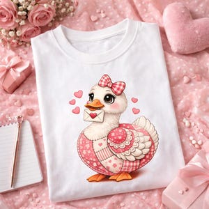 Peut inclure: T-shirt blanc avec un motif de canard de dessin animé. Le canard porte un nœud rose et tient une lettre avec un cœur. Il a un motif patchwork rose et blanc, entouré de cœurs roses. Un design charmant et romantique.