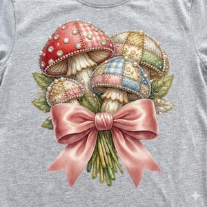 Peut inclure: T-shirt gris avec une illustration d'un bouquet de champignons. Les champignons ont des chapeaux patchwork rouges, roses et beiges, ornés de perles. Un grand nœud rose attache le bouquet, avec des tiges et des feuilles vertes.