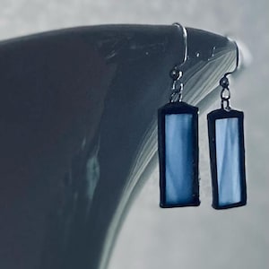 May include: A pair of rectangular earrings with a light blue center and black borders. The earrings are suspended from silver-colored ear wires. The background is a blurred, neutral color.