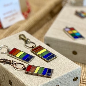 May include: Three rainbow-colored glass tile keychains with silver metal rings and clasps. The keychains are arranged on a light brown surface with a beige fabric background.