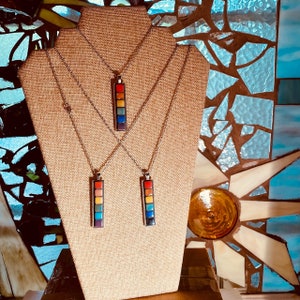 May include: Three silver necklaces with rainbow colored square tile pendants. The necklaces are displayed on a brown fabric covered mannequin.