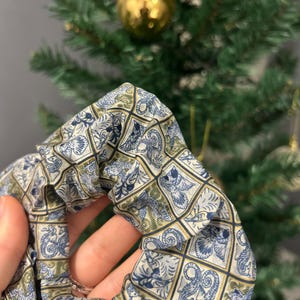 Puede incluir: Una scrunchie estampada en tonos azules, verdes y dorados. La tela presenta un diseño cuadrado repetido con ilustraciones ornamentadas y estilizadas. La scrunchie se sostiene, mostrando la textura y el patrón de la tela. Un árbol de Navidad está en el fondo.