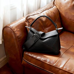 May include: A black leather handbag with a geometric design, featuring a top handle and a detachable shoulder strap. The bag is set against a brown leather sofa, with natural light from a nearby window.