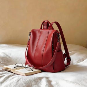 Op de afbeelding: Een rode leren rugzak met een handvat en verstelbare bandjes. De tas heeft een voorvak met rits en een zijvak. Een boek en een bril liggen voor de rugzak.