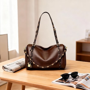 Könnte beinhalten: Eine braune Lederhandtasche mit einem oberen Griff und einem abnehmbaren Schulterriemen. Der Riemen und der Griff weisen ein sich wiederholendes goldenes Sternmuster auf. Ein kleines goldenes Schloss hängt vorne. Eine schwarze Sonnenbrille und eine Zeitschrift liegen auf einem Holztisch.