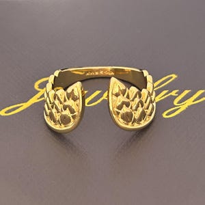 Op de afbeelding: Een goudkleurige ring met een uniek ontwerp. De ring heeft twee sierlijke, getextureerde secties die naar binnen buigen, waardoor een open ontwerp ontstaat. De ring staat tegen een donkere achtergrond.