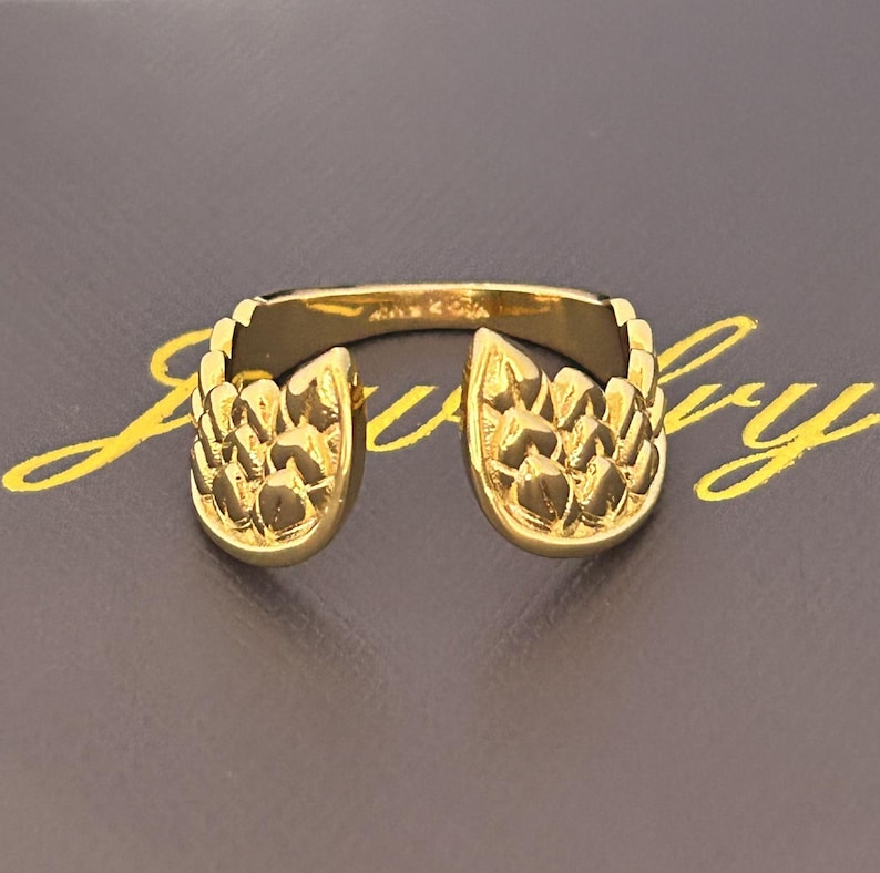 Op de afbeelding: Een goudkleurige ring met een uniek ontwerp. De ring heeft twee sierlijke, getextureerde secties die naar binnen buigen, waardoor een open ontwerp ontstaat. De ring staat tegen een donkere achtergrond.