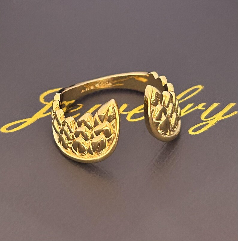 Op de afbeelding: Een goudkleurige ring met een open ontwerp. De ring heeft twee gestructureerde, bladachtige vormen aan weerszijden van de opening. De ring staat tegen een donkere achtergrond met gouden tekst.