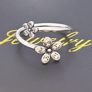 May include: A silver-toned ring featuring two daisy flower designs. Each flower has five petals and is embellished with small, clear gemstones. The ring has a curved, open design, and is set against a dark gray background.