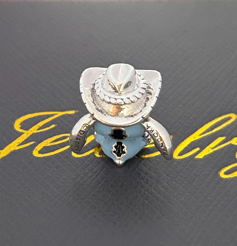 Pu&ograve; includere: Un charm d'argento raffigurante un personaggio con cappello da cowboy e abito blu. Il charm presenta le scritte "Disney" e "Pandora". &Egrave; posto su una superficie nera con scritte dorate.