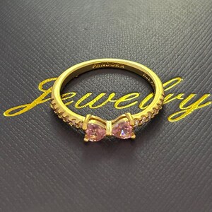 Può includere: Un anello color oro con una gemma rosa a forma di fiocco al centro. La fascia dell'anello è incastonata con piccole gemme trasparenti. La parola "PANDORA" è incisa all'interno dell'anello. L'anello è su una superficie nera con la parola "Jewelry" in oro.