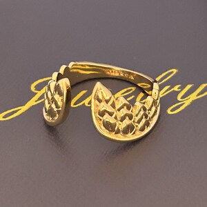 Op de afbeelding: Een goudkleurige ring met een textuurontwerp. De ring heeft een open ontwerp met een gedetailleerd, bladachtig patroon aan de voorkant en zijkant. De ring is geplaatst tegen een donkere achtergrond met gouden letters.