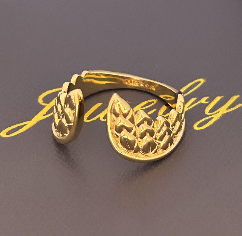 Op de afbeelding: Een goudkleurige ring met een textuurontwerp. De ring heeft een open ontwerp met een gedetailleerd, bladachtig patroon aan de voorkant en zijkant. De ring is geplaatst tegen een donkere achtergrond met gouden letters.