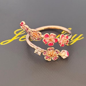 Może przedstawiać: Pierścionek z różowego złota z kwiatowym wzorem z czerwonymi i różowymi kwiatami emalii oraz małymi akcentami z czerwonych kamieni szlachetnych. Pierścionek ma delikatny, otwarty design.