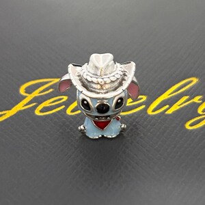 Pu&ograve; includere: Un charm blu e argento raffigurante un personaggio dei cartoni animati con un cappello da cowboy e una bandana rossa. Il charm ha occhi neri e orecchie rosa. Il charm &egrave; posto su uno sfondo scuro.