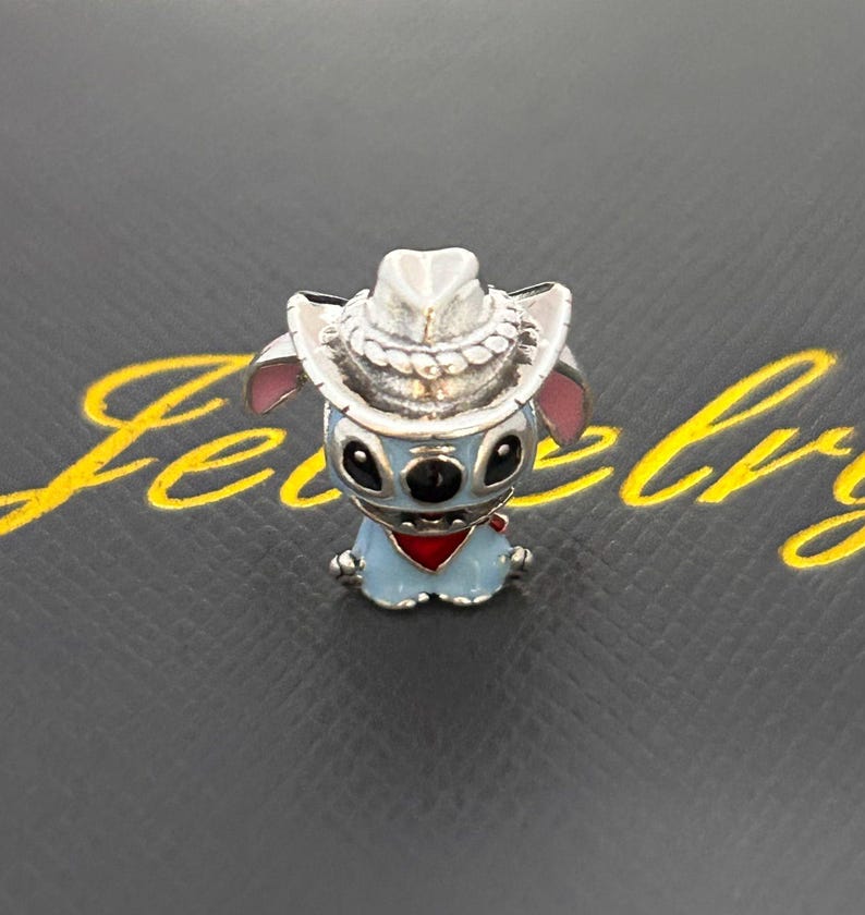 Pu&ograve; includere: Un charm blu e argento raffigurante un personaggio dei cartoni animati con un cappello da cowboy e una bandana rossa. Il charm ha occhi neri e orecchie rosa. Il charm &egrave; posto su uno sfondo scuro.