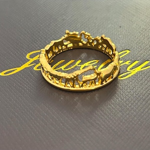 Op de afbeelding: Een goudkleurige ring met een safari-dierontwerp. De ring heeft giraffen, olifanten en andere dieren in een uitgesneden ontwerp. De ring ligt op een donkergrijze ondergrond met het woord "Jewelry" in goud.