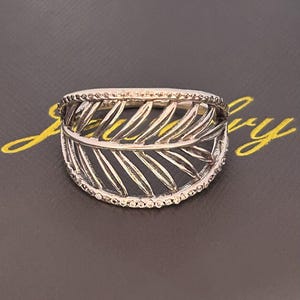 Può includere: Anello color argento con un design a foglia. L'anello presenta un motivo a foglia dettagliato al centro, circondato da una fascia di piccoli accenti scintillanti. L'anello è posto su uno sfondo scuro.