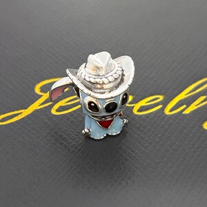 Pu&ograve; includere: Un charm argentato raffigurante un personaggio dei cartoni animati blu con un cappello da cowboy e un fazzoletto rosso. Il charm ha occhi neri e orecchie rosa. Lo sfondo &egrave; grigio scuro con scritte dorate.