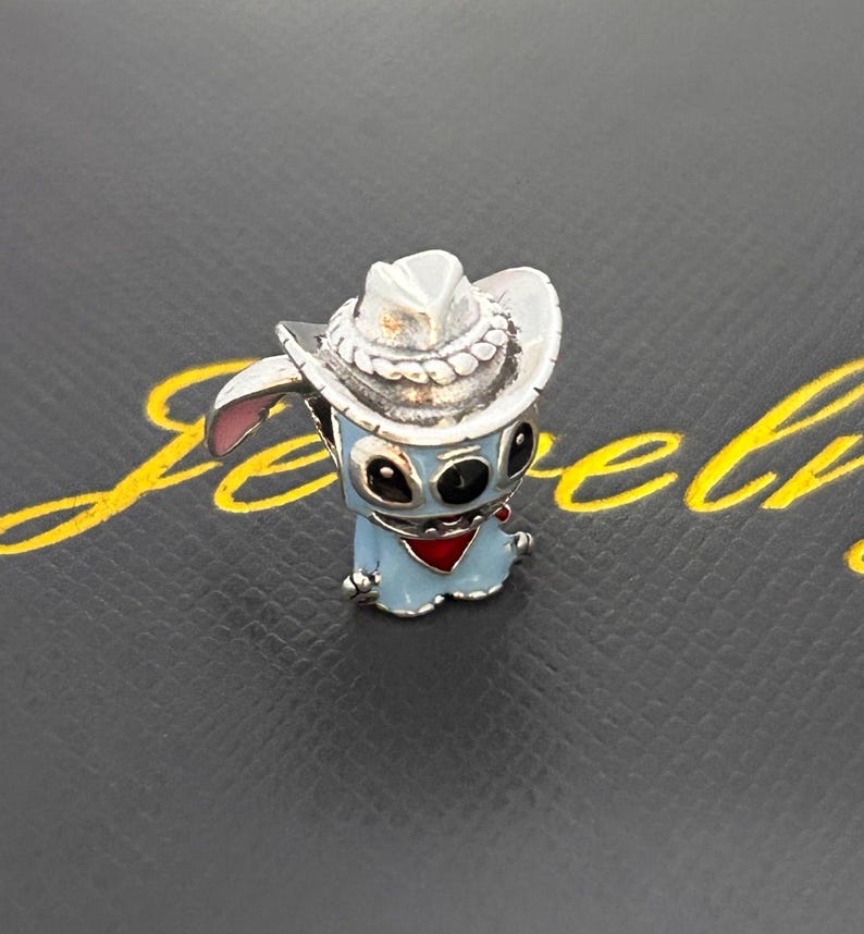 Pu&ograve; includere: Un charm argentato raffigurante un personaggio dei cartoni animati blu con un cappello da cowboy e un fazzoletto rosso. Il charm ha occhi neri e orecchie rosa. Lo sfondo &egrave; grigio scuro con scritte dorate.