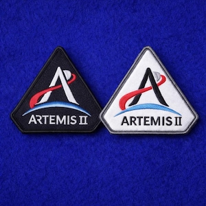 May include: Two triangular patches with the text "ARTEMIS II". One patch is black with white, red, and blue accents. The other is white with black, red, and blue accents. Both patches feature a stylized "A" design.