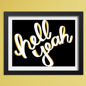 May include: A black framed print with white lettering on a black background that says "hell yeah". The print is hanging on a yellow wall between two light beige chairs with light brown wicker backs. A pink side table with a book stack and a potted plant sits between the chairs.