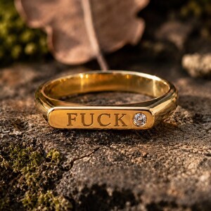 May include: A gold-colored ring with the word "FUCK" engraved on a rectangular plate. A small, clear gemstone is set on the right side of the plate. The ring is displayed on a textured, brown surface.