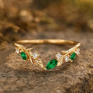 May include: A gold ring featuring a V-shaped band designed with metal leaves, three marquise-cut green emeralds, and four round-cut clear white diamonds. The band has a polished finish and follows a curved, nature-inspired contour design.
