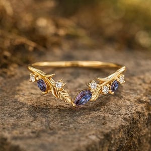Può includere: Un anello color oro con un design a foglie e gemme. L'anello presenta piccole gemme trasparenti e gemme viola più grandi a forma ovale. L'anello è posto su uno sfondo naturale e strutturato.