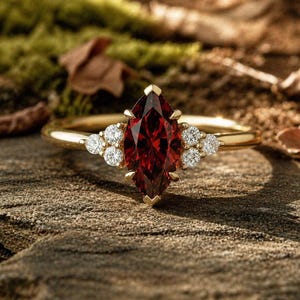 Puede incluir: Un anillo de oro con una gema roja oscura en forma de marquesa como pieza central, flanqueada por grupos de pequeñas gemas blancas redondas. El anillo se presenta sobre un fondo natural y texturizado, resaltando su elegante diseño.