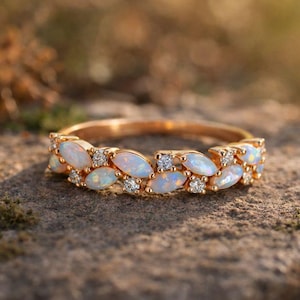 May include: A gold ring with a row of oval opals and small diamonds. The opals are milky white, and the diamonds sparkle. The ring is set on a stone surface, with a blurred background. The ring is likely made of gold.