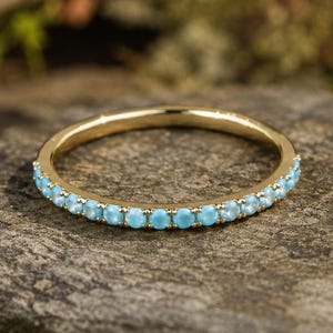 Peut inclure: Une bague dorée ornée d'une rangée de petites pierres bleues claires. La bague est posée sur une surface texturée gris-brun, avec un arrière-plan flou de feuillage.