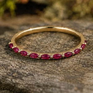 May include: A thin gold band ring featuring a series of small, marquise-cut red rubies set along the top half of the circumference. The metal has a polished finish, and the gemstones are arranged in a continuous, repeating pattern.