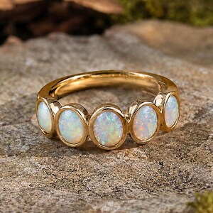 May include: A gold ring featuring five oval opals. The opals display a play of color, with flashes of white, blue, and pink. The ring is set on a stone surface with a blurred background of leaves and moss.