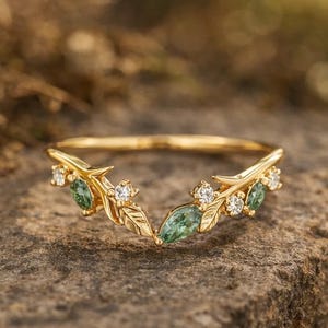 Puede incluir: Un anillo dorado con un diseño de hojas y piedras preciosas. El anillo presenta pequeñas piedras preciosas redondas y claras y piedras preciosas verdes ovaladas más grandes. El anillo está sobre una superficie de piedra.