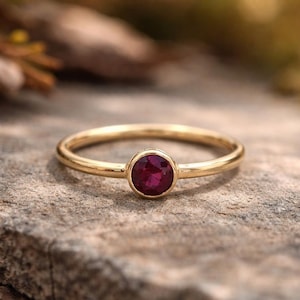 Puede incluir: Un anillo de oro con una gema redonda de color rojo intenso. La banda del anillo es delgada y pulida, y la gema está engastada en un bisel simple. El anillo se muestra sobre una superficie gris texturizada.