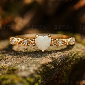 Könnte beinhalten: Ein goldener Ring mit einem herzförmigen Opal-Mittelstein. Der Ring hat kleine, klare Akzentsteine und dekorative Details am Band. Der Ring liegt auf einer natürlichen, moosigen Oberfläche.