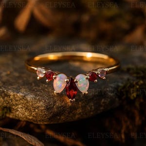 May include: A gold ring with a curved design, set with a variety of gemstones. The ring features a central cluster of pear-shaped and oval-cut red and white gemstones, with smaller red and pink stones on either side. The ring is displayed on a natural stone surface.