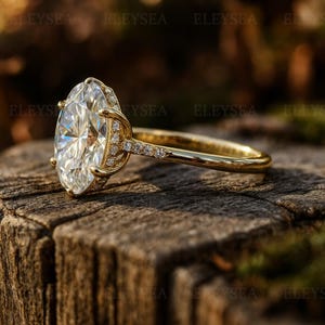 May include: A gold engagement ring with a large, oval-cut diamond as the centerpiece. The band is gold, with smaller diamonds set along the sides of the main stone and part of the band. The ring is set on a wooden surface.