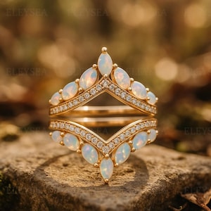 May include: A gold-colored ring set with a crown design. The ring features oval-shaped opals and small, clear gemstones. The ring is set on a stone surface.