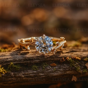 Puede incluir: Un anillo de compromiso dorado con una gema grande, redonda y transparente. La banda del anillo presenta un diseño inspirado en la naturaleza con delicados detalles de ramas y hojas. El anillo se encuentra sobre un fondo natural borroso.