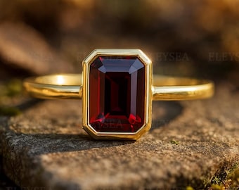 Puede incluir: Un anillo de oro con una piedra preciosa rectangular de color rojo intenso. La piedra está engastada en un bisel de oro, y la banda es de oro pulido. El anillo se muestra sobre una superficie natural y texturizada.