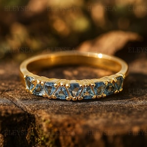 May include: A gold band ring featuring a row of triangular, light blue gemstones. The ring is set on a wooden surface, with a blurred background of green and brown tones. The ring's design is simple yet elegant.
