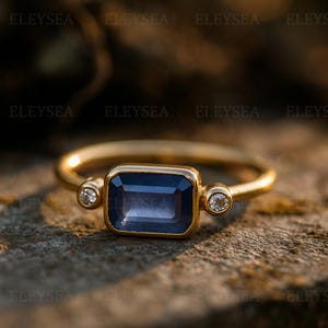 Puede incluir: Un anillo de oro con una piedra preciosa rectangular azul oscuro como pieza central, flanqueada por dos piedras preciosas redondas y claras más pequeñas. El anillo se encuentra sobre un fondo natural borroso, resaltando el diseño y la artesanía de la joya.